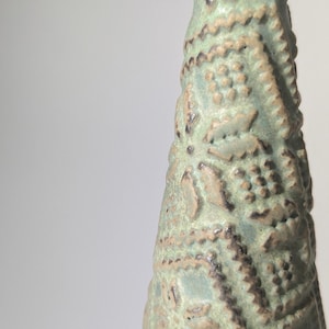 May include: A ceramic vase with a light green glaze and a detailed embossed pattern. The vase is cone-shaped and has a narrow opening.