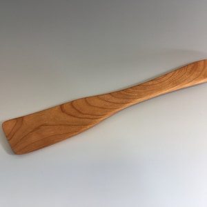May include: A wooden spatula with a natural wood finish. The spatula is long and thin with a slightly curved handle.