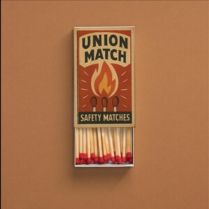 Op de afbeelding: Een rechthoekige luciferdoos met de woorden "UNION MATCH" en "SAFETY MATCHES" op de voorkant. De luciferdoos heeft een afbeelding van een vlam en drie lucifers. De doos is open en onthult talrijke houten lucifers met rode uiteinden.