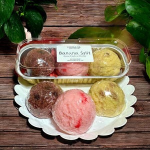 May include: A clear plastic container and a white scalloped plate hold soy wax melts. The container has three melts, and the plate has three melts. The melts are brown, pink, and yellow. The label reads "Banana Split".