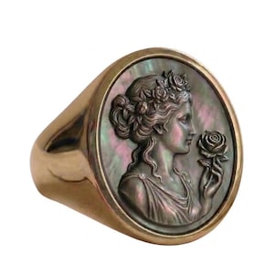 Può includere: Un anello a sigillo color oro con un rilievo dettagliato di una donna che tiene una rosa. La faccia dell'anello ha uno sfondo in madreperla e il profilo della donna è finemente scolpito. L'anello ha un design classico ed elegante.
