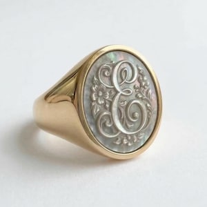 Può includere: Un anello sigillo in oro con un inserto ovale in madreperla. Al centro dell'inserto è incisa una lettera maiuscola E ornata, circondata da delicati motivi floreali e volute, il tutto montato su una fascia in oro lucido.