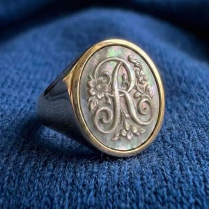 Puede incluir: Anillo de plata con bisel dorado, con una cara ovalada de nácar. La cara está grabada con la letra "R" en un diseño floral ornamentado. El anillo está sobre un fondo azul.