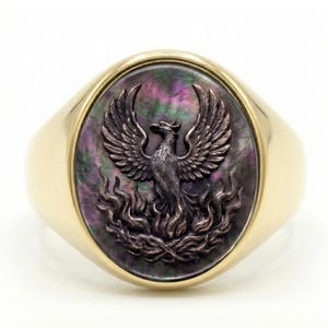 May include: A gold signet ring featuring an oval inlay of iridescent dark mother-of-pearl. Centered on the inlay is a raised, metallic relief of a phoenix with wings spread wide, rising from stylized flames that curve along the bottom edge.