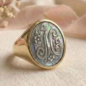 Puede incluir: Un anillo de sello dorado con una cara de nácar. La cara presenta un diseño floral y la letra "M". El anillo tiene un acabado pulido y un estilo clásico y elegante.