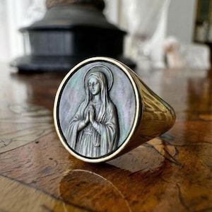 Könnte beinhalten: Ein goldfarbener Ring mit einem detaillierten Relief einer religiösen Figur mit gefalteten Händen. Die Ringoberfläche hat einen dunklen, schillernden Hintergrund, und das Band ist aus poliertem Gold. Der Ring liegt auf einer Holzoberfläche.