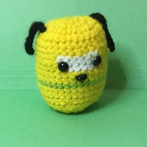 May include: A handmade, yellow crocheted creature with black ears, black eyes, and a white and green stripe. The amigurumi has a rounded shape and is set against a green background.