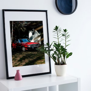 以下が含まれることがあります： 白い建物の前に駐車された赤いヴィンテージカーが写ったフレームプリント。車が焦点で、ヘッドライトやグリルなどの詳細が見えます。プリントは、ピンクの花瓶と鉢植えの植物が置かれた白い棚にあります。