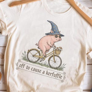 Könnte beinhalten: Cremefarbenes T-Shirt mit einer Illustration eines Schweins auf einem Fahrrad, das einen Zaubererhut und eine Sonnenbrille trägt. Der Text "off to cause a kerfuffle" steht darunter. Das Shirt hängt an einem Holzkleiderbügel.