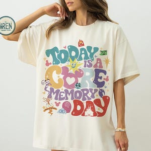 May include: A cream-coloured t-shirt featuring the phrase "Today is a Core Memory Day" in a vibrant, retro font. The design incorporates cartoon characters and a castle illustration, giving a playful and nostalgic feel.