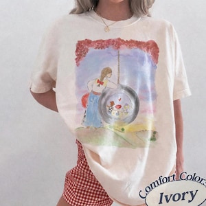 May include: An ivory short-sleeve t-shirt featuring a watercolor-style graphic of a person with braided hair standing next to a tire swing. The illustration includes a sunset sky, red foliage, and small figures inside the tire. The shirt has a crew neck.