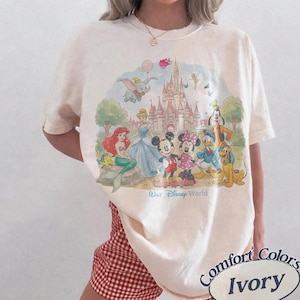 May include: An ivory short-sleeve t-shirt featuring a colorful graphic print of Disney characters including Mickey Mouse, Minnie Mouse, Ariel, Cinderella, Dumbo, and Goofy in front of a castle with the text Walt Disney World printed below.