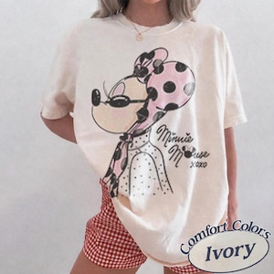 May include: Ivory-colored t-shirt featuring a Minnie Mouse graphic with a pink polka dot bow and scarf. The text "Minnie Mouse xoxo" is below the graphic. The shirt is paired with red and white checkered shorts.