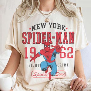 May include: Cream-colored t-shirt featuring a graphic of Spider-Man in red and blue, with the text "NEW YORK SPIDER-MAN 1962 FIGHT CRIME Spidey Sense". The design has a vintage, distressed look.
