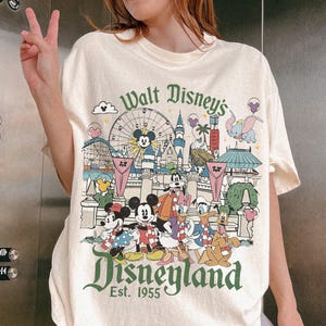 May include: Cream-colored t-shirt featuring a Disneyland graphic with Mickey Mouse, Minnie Mouse, Donald Duck, and other Disney characters. The design includes a Ferris wheel, castle, and the text "Walt Disney's Disneyland Est. 1955."