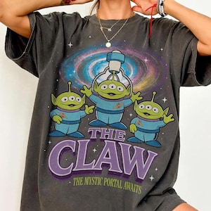以下が含まれることがあります： 青い衣装を着た3人の緑色のエイリアンのグラフィックと「THE CLAW」の文字、その下に「THE MYSTIC PORTAL AWAITS」が描かれた濃い灰色のTシャツ。背景にはカラフルな銀河のデザインが描かれています。