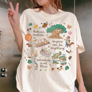 May include: Cream-colored t-shirt featuring colorful cartoon illustrations and text. The shirt includes phrases like "Hakuna Matata" and "Adventure is out there." Various characters and icons are depicted, including a tree, animals, and a safari vehicle.