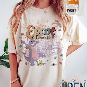 May include: A white t-shirt with a colorful graphic of Figment, the purple dragon from Epcot's Imagination! pavilion, in front of the Spaceship Earth icon. The text "Epcot Tour 1928" is above Figment.
