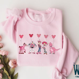 Może przedstawiać: R&oacute;żowa bluza z nadrukiem postaci z Toy Story trzymających r&oacute;żowe balony w kształcie serca. Postacie to Woody, Buzz Astral, Jessie, Bulterier i Rex.