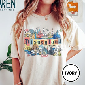 May include: A cream colored t-shirt with a colorful graphic of Disneyland Resort. The graphic includes iconic attractions like Sleeping Beauty's Castle, the Matterhorn, and the Haunted Mansion. The text "Disneyland Resort" is printed in red and yellow.