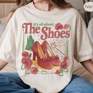 May include: Ivory-colored t-shirt with a graphic that reads "It's all about The Shoes." The graphic features red high heels, flowers, and a star wand. The shirt is a relaxed fit.