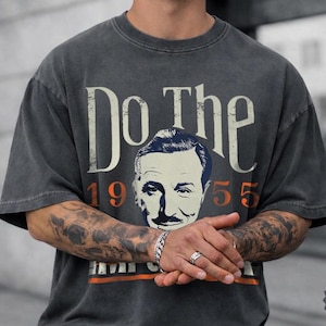 May include: Pepper-colored t-shirt with the text "Do The" above a vintage portrait and the number "1955". The shirt has a distressed look, with a faded design and a crew neck.
