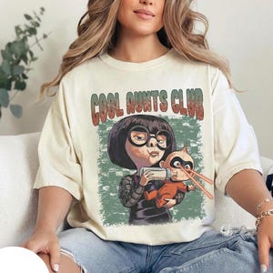 Può includere: T-shirt color avorio con una grafica di un personaggio dei cartoni animati che tiene in braccio un bambino con raggi laser. Il testo "COOL AUNTS CLUB" è sopra la grafica. La maglietta è indossata da una persona in jeans blu.