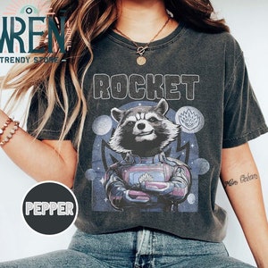 Puede incluir: Una camiseta gris oscuro con un gráfico de Rocket Raccoon de Guardianes de la Galaxia. El gráfico es de estilo vintage con una combinación de colores azul, morado y blanco. El texto "ROCKET" está impreso en blanco sobre el gráfico.