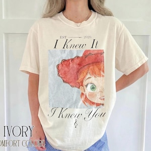 May include: An ivory short-sleeve t-shirt featuring a graphic print of a character with red hair and a red hat. The text reads "EST 2026", "I Knew It", and "I Knew You" in a black serif font. The design is centered on the chest of the garment.