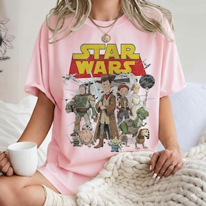 May include: A light pink t-shirt featuring a graphic of Star Wars characters in a cartoon style. The Star Wars logo is in yellow and red. Characters include Woody, Buzz Lightyear, and others. The shirt has short sleeves.