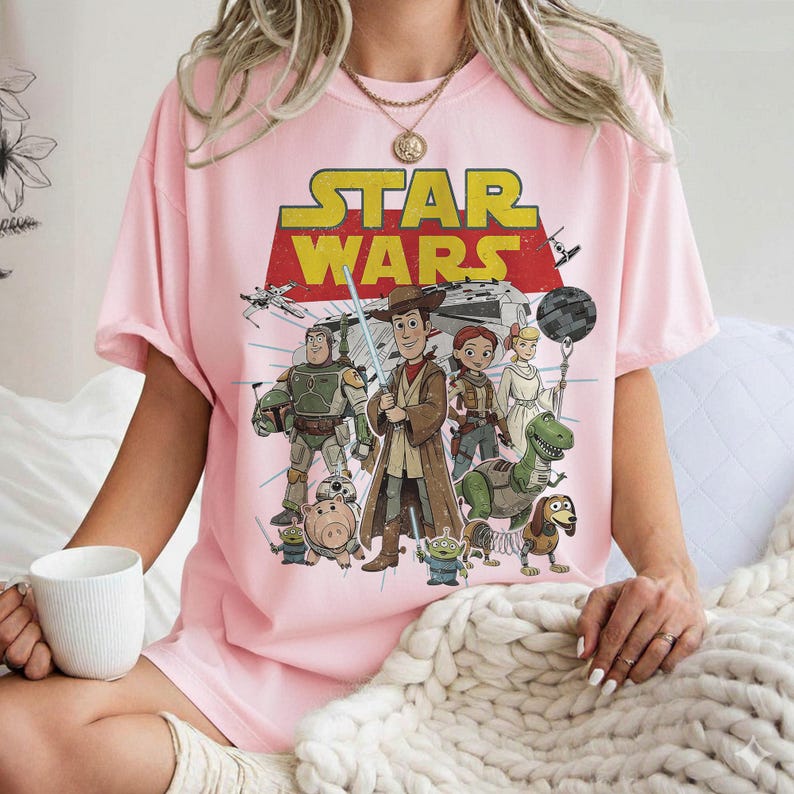May include: A light pink t-shirt featuring a graphic of Star Wars characters in a cartoon style. The Star Wars logo is in yellow and red. Characters include Woody, Buzz Lightyear, and others. The shirt has short sleeves.