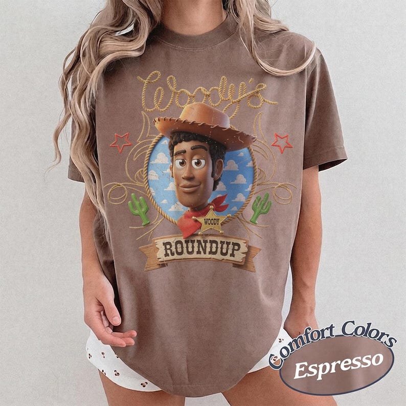 K&ouml;nnte beinhalten: Ein braunes Kurzarm-T-Shirt mit einem Aufdruck der Figur Woody aus Toy Story. Das Design zeigt einen Seilrahmen, einen Sheriffstern, gr&uuml;ne Kakteen und den Text Woody's ROUNDUP. Das T-Shirt hat einen Rundhalsausschnitt und eine lockere Passform.
