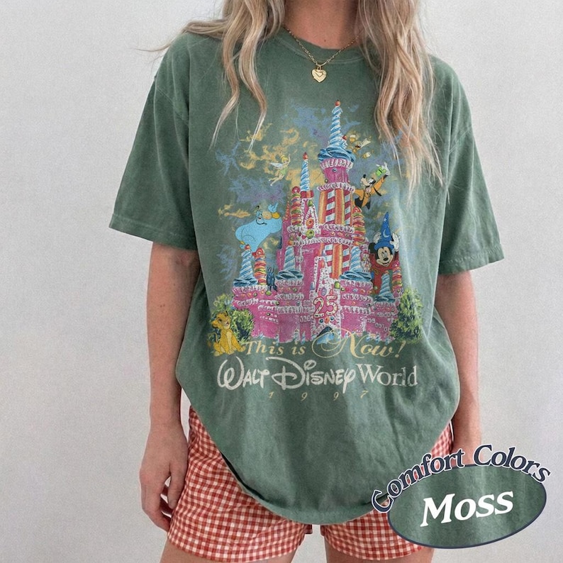 Może przedstawiać: Zielona koszulka w kolorze mchu z kolorową grafiką zamku Kopciuszka, Myszki Miki i innych postaci Disneya. Tekst "This is Now! Walt Disney World 1997" jest wydrukowany poniżej. Koszulka jest z Comfort Colors.