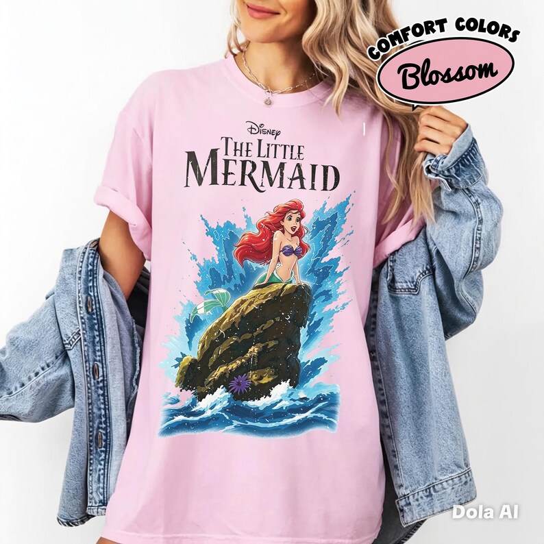 May include: A light pink short-sleeve t-shirt featuring a graphic of Ariel from The Little Mermaid sitting on a rock amidst blue ocean waves. The text reads Disney The Little Mermaid above the character. The shirt is worn under a denim jacket.