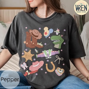 May include: Dark gray t-shirt with a colorful cartoon graphic featuring cowboy boots, a sheriff badge, a hat, a horseshoe, and a dinosaur. The shirt also has the text "Pepper" at the bottom.