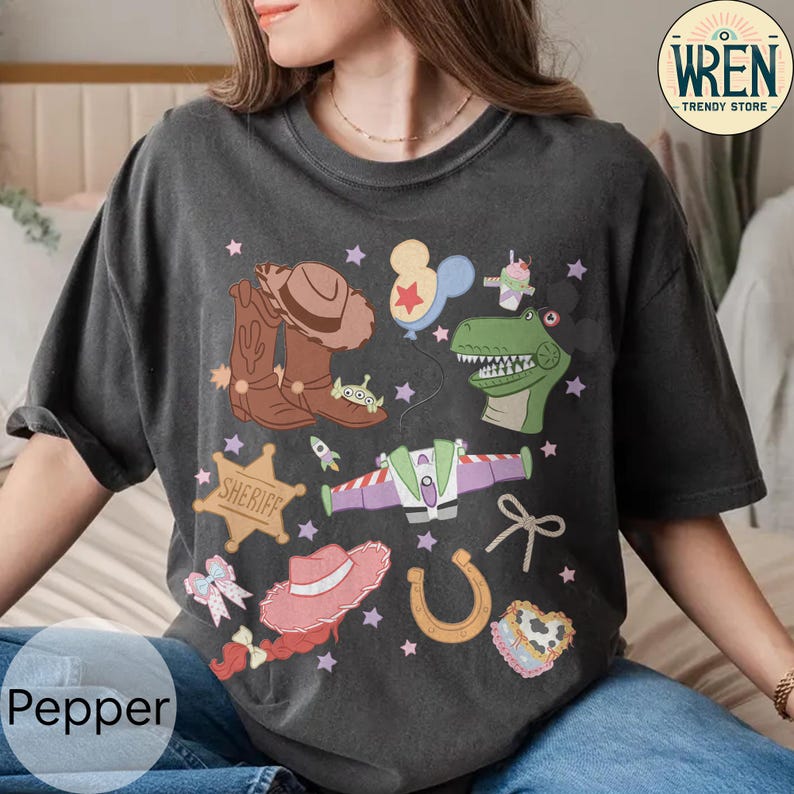 May include: Dark gray t-shirt with a colorful cartoon graphic featuring cowboy boots, a sheriff badge, a hat, a horseshoe, and a dinosaur. The shirt also has the text "Pepper" at the bottom.