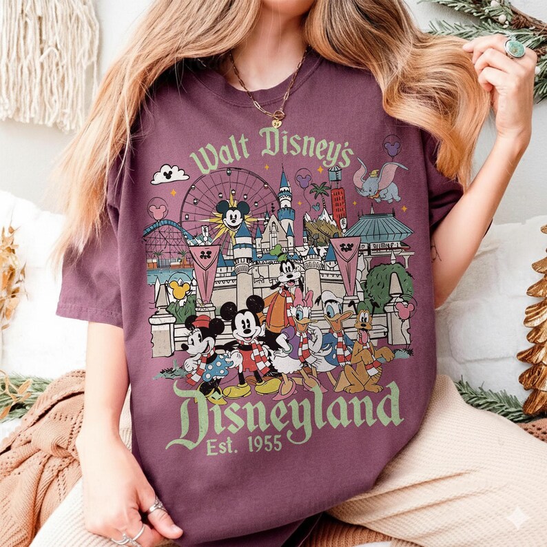 May include: A maroon t-shirt featuring a colorful graphic of Disneyland with Mickey Mouse, Minnie Mouse, Donald Duck, and other Disney characters. The text "Walt Disney's Disneyland" and "Est. 1955" are also displayed.