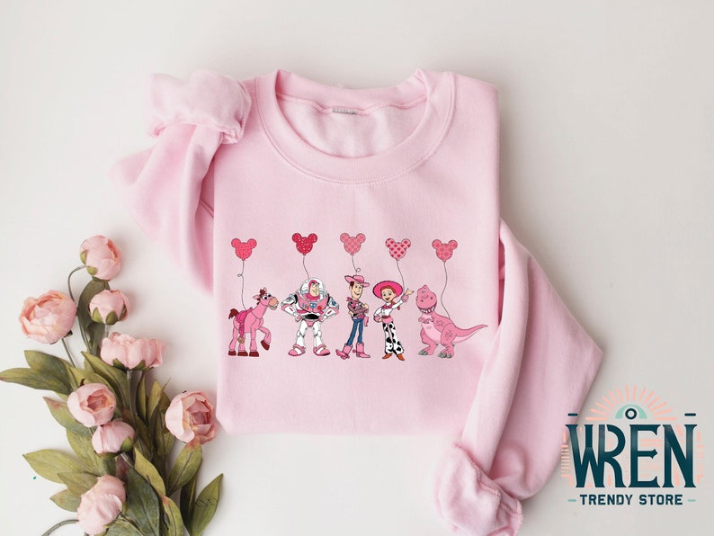 Może przedstawiać: R&oacute;żowa bluza z nadrukiem postaci z Toy Story trzymających r&oacute;żowe balony w kształcie serca. Postacie to Woody, Buzz Astral, Jessie, Bulterier i Rex.