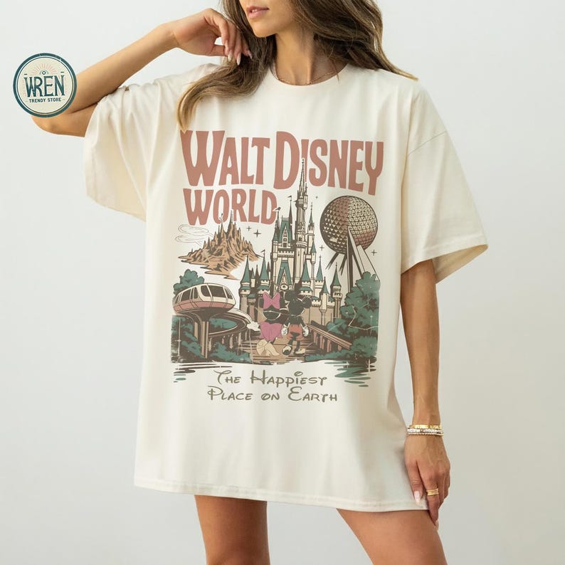 Może przedstawiać: Kremowa koszulka z grafiką w stylu vintage z Walt Disney World. Projekt zawiera napisy "Walt Disney World", zamek, Spaceship Earth i frazę "The Happiest Place on Earth."