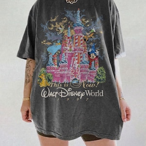 Może przedstawiać: Ciemnoszara koszulka z kolorową grafiką zamku Kopciuszka i postaciami Disneya. Tekst "This is Now! Walt Disney World 1997" jest wydrukowany poniżej. Koszulka ma kr&oacute;tkie rękawy i luźny kr&oacute;j.