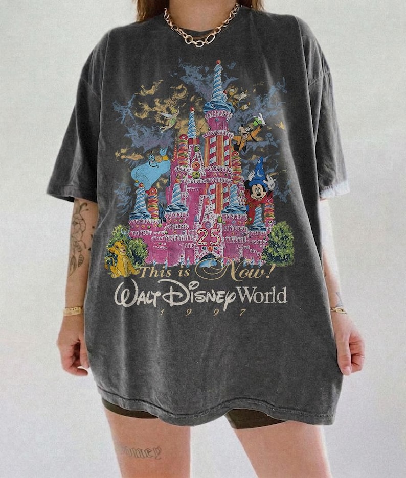 Może przedstawiać: Ciemnoszara koszulka z kolorową grafiką zamku Kopciuszka i postaciami Disneya. Tekst "This is Now! Walt Disney World 1997" jest wydrukowany poniżej. Koszulka ma kr&oacute;tkie rękawy i luźny kr&oacute;j.