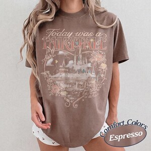 Puede incluir: Una camiseta marr&oacute;n de manga corta con un estampado gr&aacute;fico que incluye el texto Today was a FAIRY TALE y Can you feel this magic in the air? El dise&ntilde;o presenta una ilustraci&oacute;n de un castillo, mariposas y elementos florales.