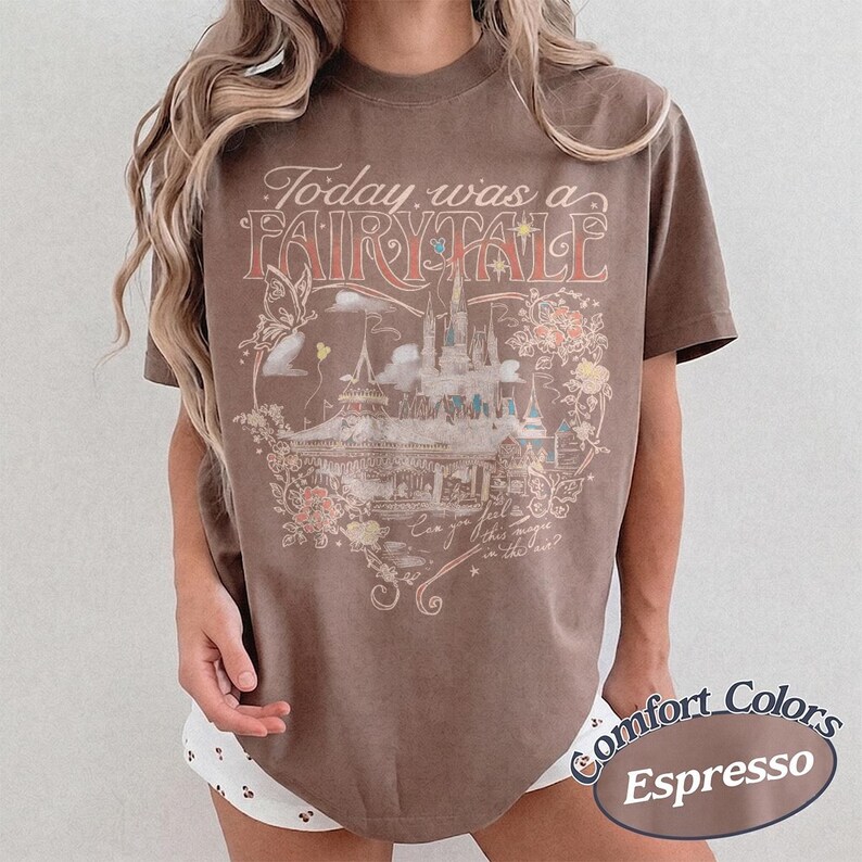 Puede incluir: Una camiseta marr&oacute;n de manga corta con un estampado gr&aacute;fico que incluye el texto Today was a FAIRY TALE y Can you feel this magic in the air? El dise&ntilde;o presenta una ilustraci&oacute;n de un castillo, mariposas y elementos florales.
