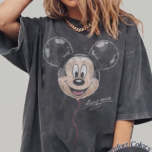 Op de afbeelding: Donkergrijze t-shirt met een verweerde Mickey Mouse ballonafbeelding. Het ontwerp bevat de tekst "Mickey Mouse" in script. Het shirt is gelabeld "Comfort Colors Pepper".