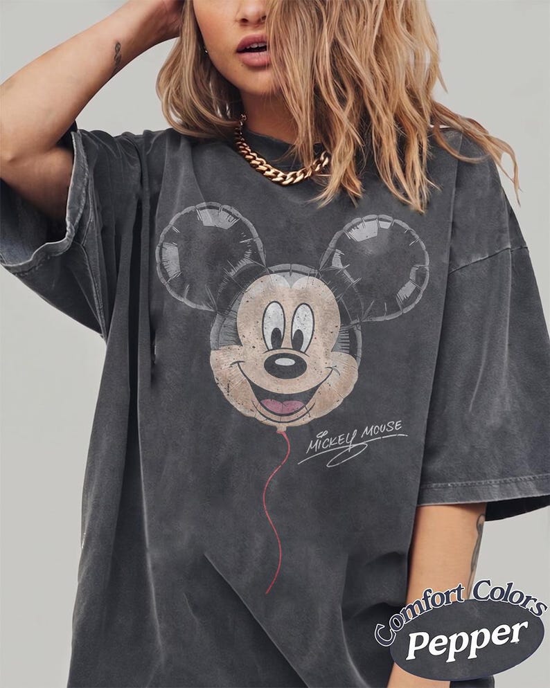 Op de afbeelding: Donkergrijze t-shirt met een verweerde Mickey Mouse ballonafbeelding. Het ontwerp bevat de tekst "Mickey Mouse" in script. Het shirt is gelabeld "Comfort Colors Pepper".