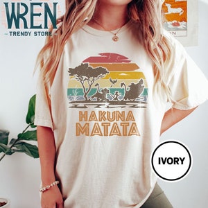 May include: A white t-shirt with a vintage-style sunset graphic featuring the Lion King characters Simba, Timon, and Pumbaa. The text "Hakuna Matata" is printed below the graphic. The shirt is labeled "Ivory".