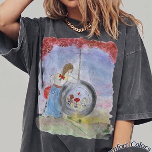 May include: A charcoal gray short-sleeve t-shirt featuring a watercolor-style graphic of a person with a braid leaning against a tire swing. Inside the tire, three small figures are visible. The text Comfort Colors Pepper is printed in the bottom corner.