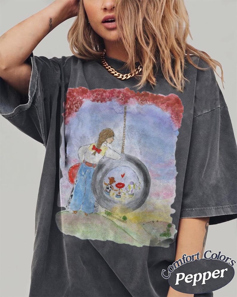 May include: A charcoal gray short-sleeve t-shirt featuring a watercolor-style graphic of a person with a braid leaning against a tire swing. Inside the tire, three small figures are visible. The text Comfort Colors Pepper is printed in the bottom corner.