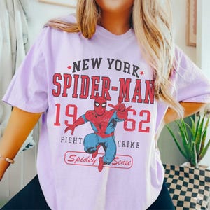 Op de afbeelding: Lichtpaars T-shirt met een rood en blauw Spider-Man-ontwerp en de tekst "NEW YORK SPIDER-MAN 1962 FIGHT CRIME" in het rood. De tekst "Spidey Sense" staat in een rood kader.