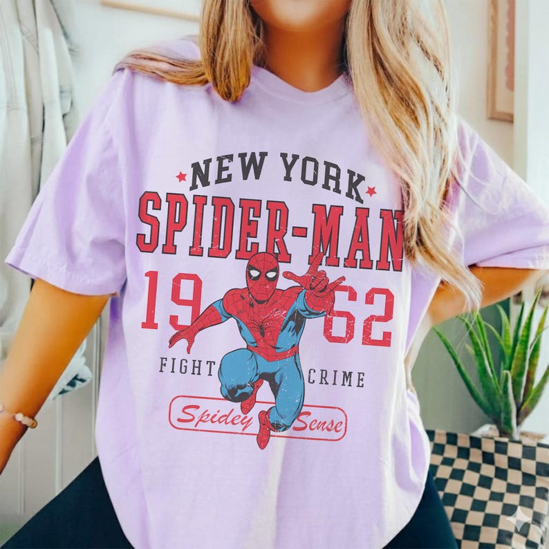 Op de afbeelding: Lichtpaars T-shirt met een rood en blauw Spider-Man-ontwerp en de tekst "NEW YORK SPIDER-MAN 1962 FIGHT CRIME" in het rood. De tekst "Spidey Sense" staat in een rood kader.